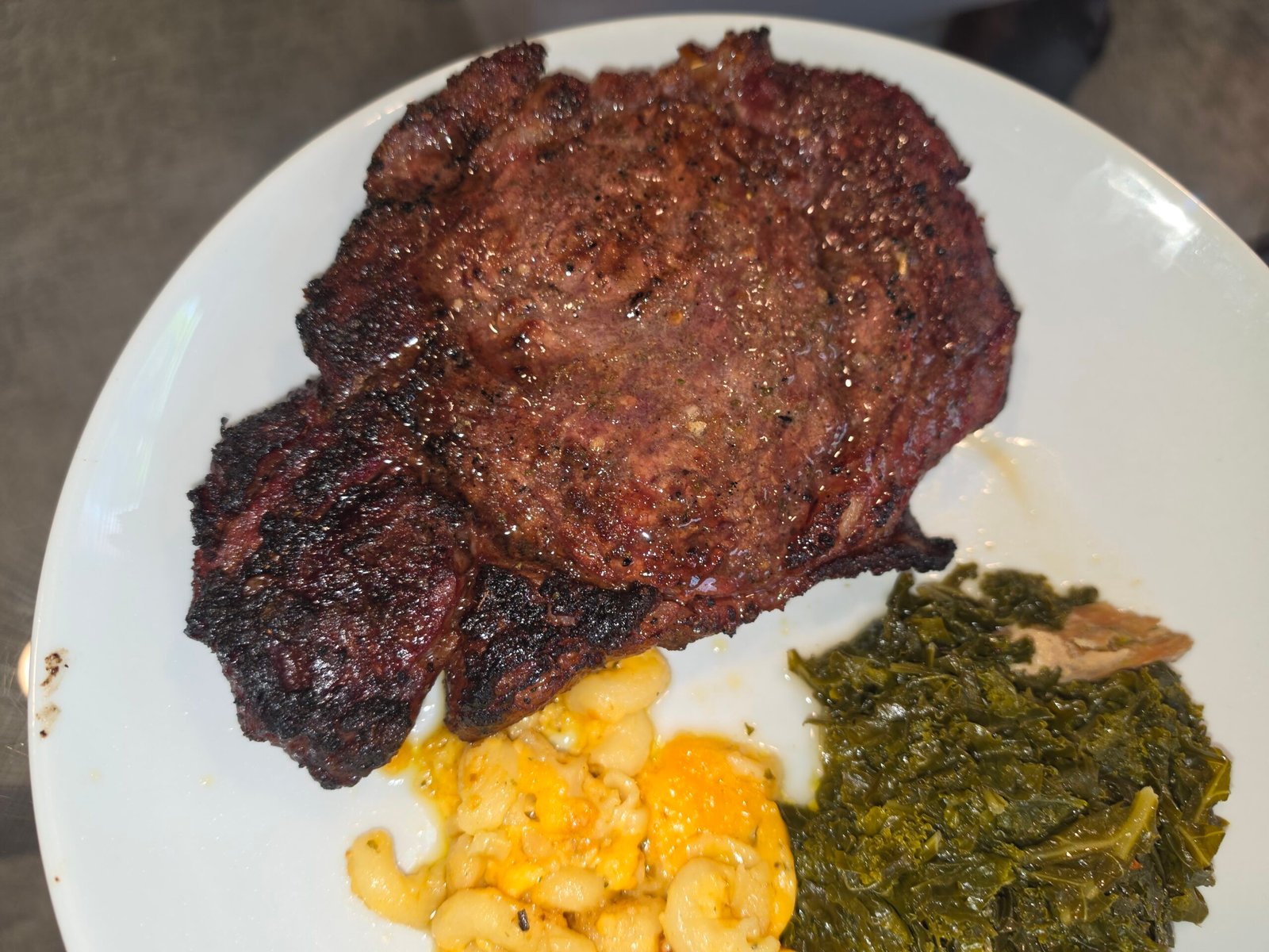 A large cooked steak served on a white plate with a side of macaroni and cheese and collard greens