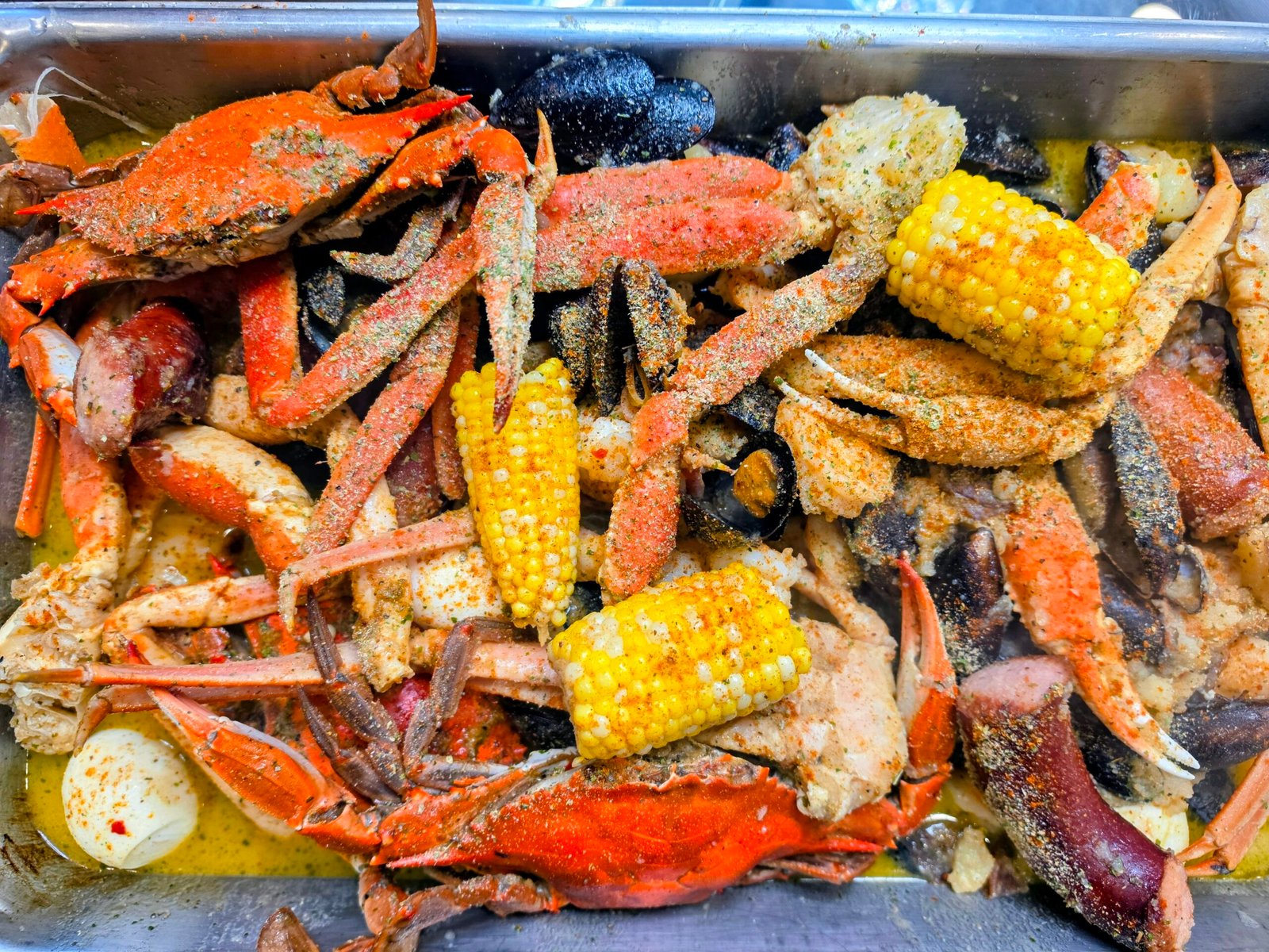 A metal tray filled with seafood boil, including blue crabs, snow crab legs, mussels, corn on the cob, and sausage pieces, all seasoned with spices and butter sauce